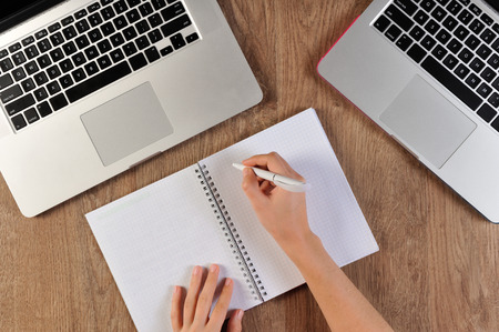 person writing in notebook infront of two laptopsの写真素材