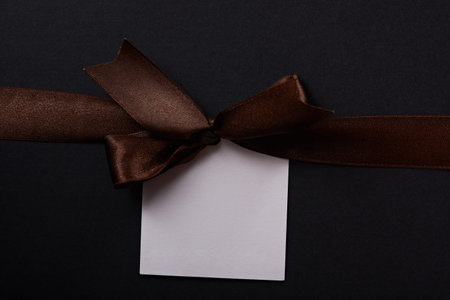 Brown gift bow with paper white label tag on dark background. Brown ribbon with empty tagの写真素材
