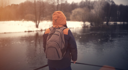 Traveler with backpack look ahead. Traveler in warm clothes on winter background. Travel concept in retro tones. One man on riverside look at water.の写真素材