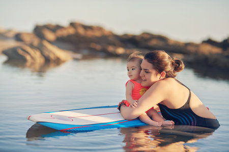 Young mom with baby on surf board in blurred ocean backgroundの写真素材