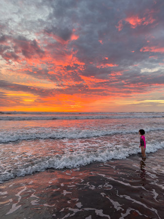 Young child stands in shallow water, enjoying waves as stunning sunset paints sky with vibrant colors.の写真素材
