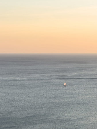 Small sailing boat navigates serene ocean waters as sun sets, casting warm hues in sky.の写真素材