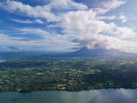 Breathtaking aerial perspective captures verdant hills and majestic volcano surrounded by ocean under dynamic clouds.の写真素材