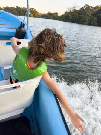 Young child in green life jacket leans out of boat, delighting in breeze as it glides over the water.の写真素材