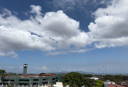 Fluffy clouds float across bright blue sky, casting shadows over urban area and distant mountains.の写真素材