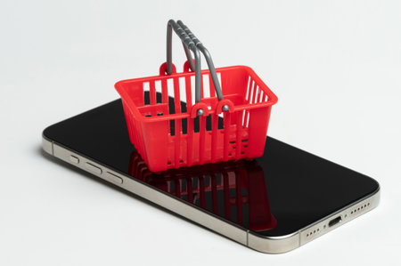 Small, red shopping basket rests on top of modern mobile phone. Background plain white, emphasizing objects.の写真素材