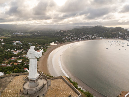 A large statue stands on a hill, offering a panoramic view of the bay below during sunset, with boats dotting the water.の写真素材