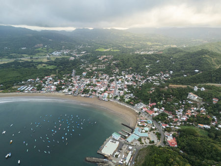 Aerial view of a coastal town showing colorful buildings, boats on the water, and lush green hills under a cloudy sky.の写真素材