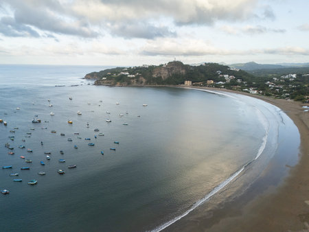 Calm waters mirror the sky as fishing boats line the shore of a coastal village at sunset, framed by green hills.の写真素材