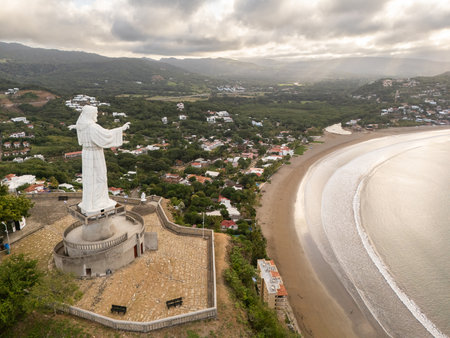 A tall statue stands on a hilltop, watching over a beachside town as golden sunlight spills across the ocean during sunset.の写真素材