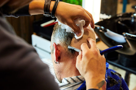 Portrait of young bald bearded man getting shaved with straight edge razor by hairdresser at...