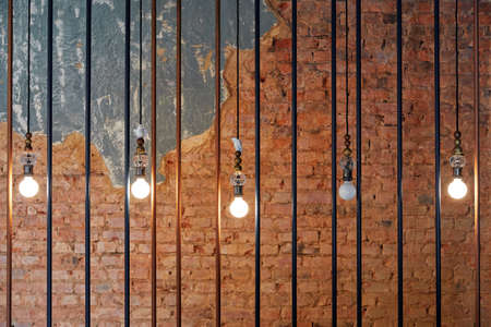 burning light bulbs, hanging in a row, behind iron bars, against the background of a brick and stone wallの写真素材