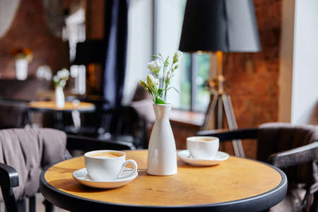 a table in a cafe on which there are two white cups of coffee and a vase with a bouquet of flowers, restaurant interiorの写真素材