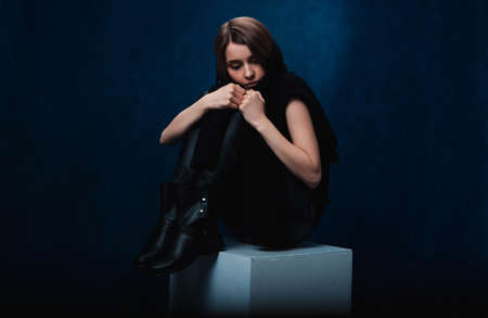 sad girl in black clothes, legs crossed, sits on a white blockの写真素材