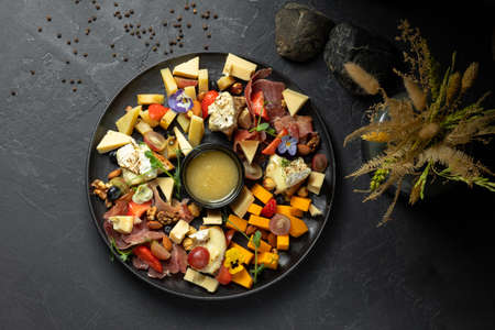 different types of cheese with fruits, nuts and meat with sauce on a black plateの写真素材