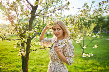 a girl with blonde hair in a flower dress stands next to a flowering treeの写真素材