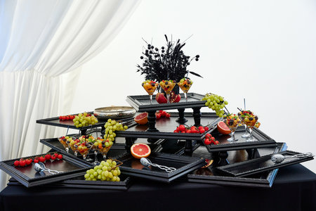 Decorated trays with fresh organic fruits put on black table in restaurant premise. Diversity of healthy food served for guests against white curtain closeupの写真素材