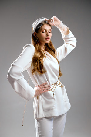 Young model in stylish outfit stands in elegant pose in grey studio. Woman with languid look enjoys professional photoshoot for fashion magazineの写真素材