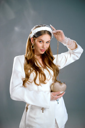 Young female model demonstrates promoting stylish handbag in shape of heart in studio. Woman in white jacket and headband poses in grey studioの写真素材