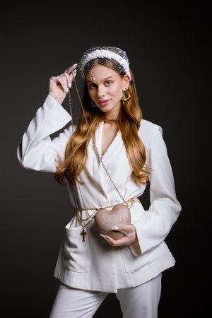 Young woman in stylish white outfit holds heart shaped handbag looking in camera. Model with long loose hair stands in studio posing for photo on black backgroundの写真素材