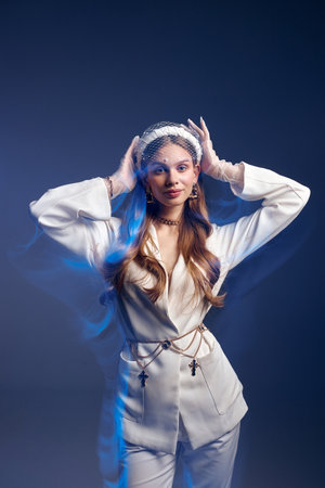 Young woman decorated with light transparent blue veil puts hands on head posing for photo in studio. Stylish model stands at dark wall illuminated with neon lightの写真素材