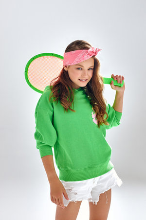 Cute teen girl in green hoodie poses for photo holding tennis racquet. Active child with pink headband stands smiling on studio white backgroundの写真素材