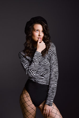 Elegant dreamy woman puts finger on lip posing for photoshoot. Young model in jumper with zebra pattern stands on dark grey background in studioの写真素材