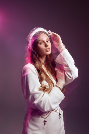 Young female model poses for photo raising hand in studio illuminated with pink neon light. Woman in white outfit looks in camera smilingの写真素材