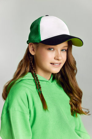Girl in baseball cap and green hoodie looks in camera smiling. Portrait of teen model with long brown hair posing in studio on grey background closeupの写真素材