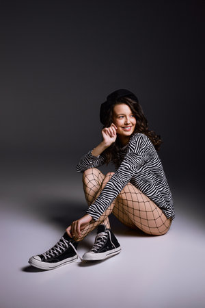 Cheerful woman in fishnet tights sits on floor in studio smiling. Young brunette model enjoys posing for photoshoot on dark grey backgroundの写真素材