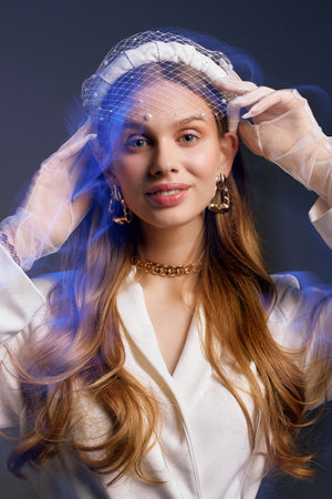 Young long-haired woman with veil looks in camera smiling. Female model poses for photo on grey background illuminated by blue neon light closeupの写真素材