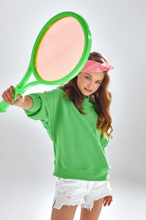 Teen girl with pink headband shows tennis racquet standing in studio. Sportive teenager in green hoodie poses for photo on white background closeupの写真素材