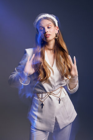 Talented female model with languid look poses for professional photoshoot in studio. Young woman in white jacket stands at dark wall illuminated by neon blue lightの写真素材