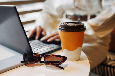 Businesswoman works on laptop and drinks coffee from cardboard cup. Lady in formal clothes puts glasses and cup with beverage on table in cafeの写真素材