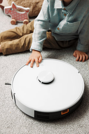 Kid boy explores and touches robot vacuum cleaner in apartment. Modern device helps owners to take care of house and get rid of dust on floorの写真素材