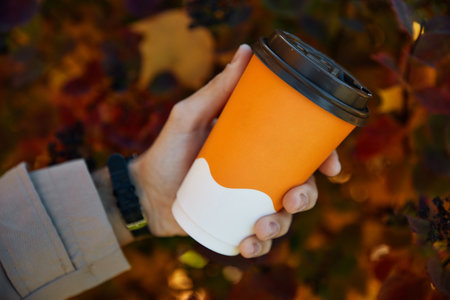 Person holds yellow cup of coffee and enjoys walking in autumn park. Tourist enjoys relaxing in autumn park against falling red leavesの写真素材