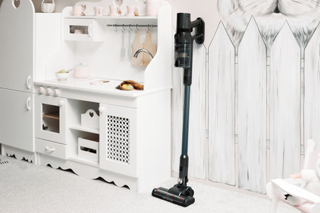Handheld wireless vacuum cleaner with removable battery for quick and accessible cleaning floors. Black vacuum cleaner stands near toy kitchenの写真素材