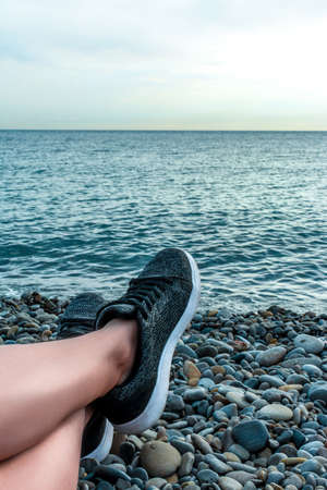 Holidays at sea, vacation and travel concept-young girl resting lying on the sea, legs in sneakers close-up, summer adventures, pebble beach, seascape copy spaseの写真素材