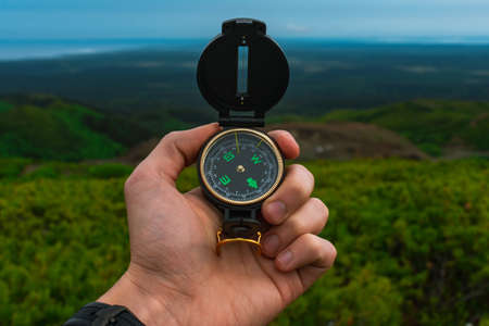 Travel, camping, orienteering and navigation concept - black magnetic compass close-up in a man s hand, blurred landscape backgroundの写真素材