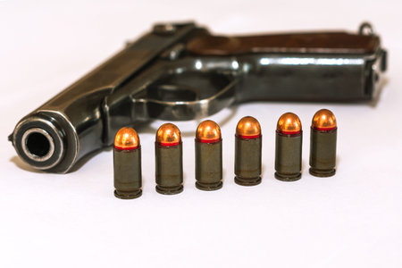 Six combat pistol cartridges of calliber 9 mm stand in front of a Makarov pistol on a white...