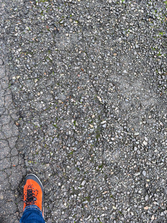 The rough asphalt surface is covered in small stones, and a person's foot in an orange shoe is...