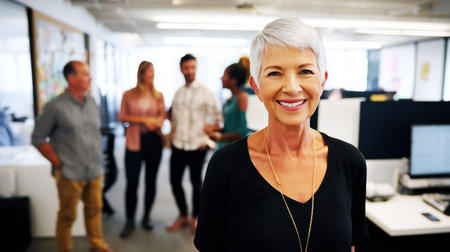 Portrait of smiling senior businesswoman in office with colleagues in the backgroundの素材