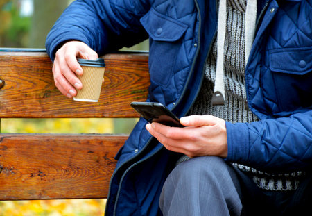 a man sits on a bench in an autumn park, holds a cup of coffee and a smartphone in his handsの写真素材