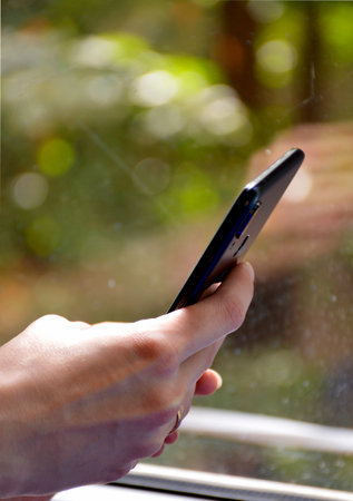 against the background of the window, female hands hold a smartphone of black colorの写真素材