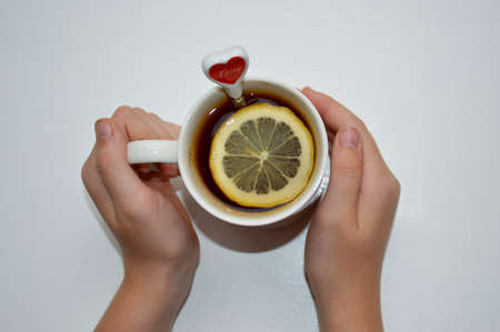female hands hold a cup of tea in which a lemon slice floats, the top of a teaspoon sticks out of the cupの写真素材
