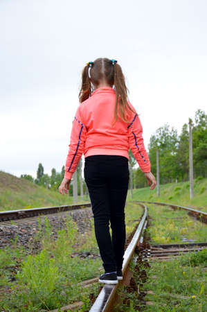 A little girl walks along the rails and balances on the railway track with her arms outstretched. Shot from the back, no face, bright sunny day in natureの写真素材