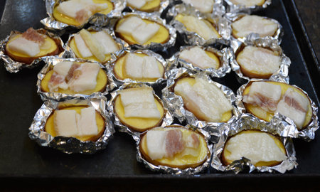 potato halves prepared for baking in the oven wrapped in foil and pieces of lard or bacon on top. preparing a hearty mealの写真素材