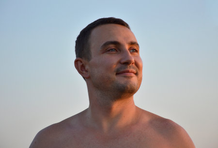 handsome man in the rays of the setting sun looks away and smiles. portrait of a young man. rest and walks in the open air on warm summer daysの写真素材