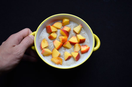 oatmeal cooked in milk with pieces of ripe peach in a bowl on a black background. tasty and healthy food for children and adults. vitamins from all fresh fruits and fiber in porridge. dietary foodの写真素材
