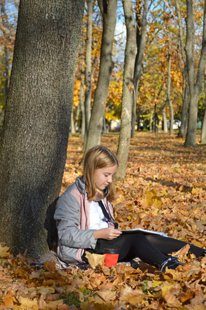 a beautiful girl sits on fallen leaves leaning against a tree trunk in an autumn park. holding a...
